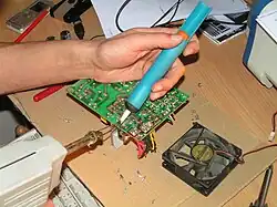 Zdjęcie przedstawia odlutowywanie elementu elektroniki. Ręka trzyma odsysacz przyłożony do płytki elektronicznej, a do tego samego punktu przykładana jest też lutownica.