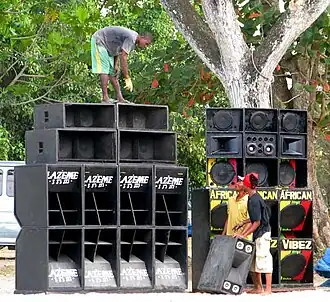 Zdjęcie z 2009 roku. Czarnoskóry mężczyzna stoi na wysokich kolumnach głośnikowych. Rozmawia on z dwoma czarnoskórymi mężczyznami, którzy trzymają na dole jeden z dużych głośników.