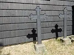 Zdjęcie wykonane w słoneczny dzień przedstawia dwa żeliwne krzyże na niewysokich postumentach. Zaraz za nimi znajduje się ściana kościoła pokryta gontem. Na ścianie widoczne wyraźne cienie krzyży.