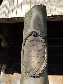 Zdjęcie przedstawia fragment złamanej kamiennej kolumny, Na kolumnie wyrzeźbiony jest wieniec, w środku którego znajduje się napis. Za kolumną znajduje się drewniana ściana kościoła i fragment gontowego daszku.
