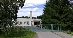 Zdjęcie przedstawia budynek szkoły podstawowej nr 11 na Chyszowie. Na pierwszym planie widać biało-szary budynek, z kilkoma oknami, zadaszonym wejściem i białym wysokim kominem. Przed budynkiem stoi ogrodzenie i brama pomalowane na zielono. Po prawej stronie, od bramy, ciągnie się chodnik, wzdłuż którego rosną sięgające ziemi iglaste drzewa. W tle zdjęcia, za szkołą widać więcej wysokich drzew.