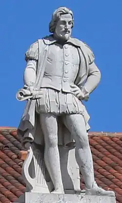 Marmurowy posąg przedstawiający człowieka. Jedną ręką trzyma zwinięte karty papieru, drugą podpiera swój bok. Ubrany jest w strój adekwatny do epoki renesansu.