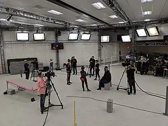 A bright white stage with actors in black suits and camera operators