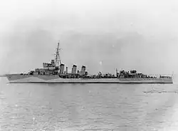 Widok na lewą burtę okrętu, stanowiska artylerii głównej i pom-pom skierowane na lewą burtę. Geometryczne figury kamuflażu na burcie rozcinają sylwetkę okrętu – jasne plamy niżej, ciemniejsze wyżej (zdjęcie czarno-białe).