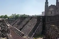 Zdjęcie kolorowe przedstawia dolne trzy kondygnacje zbudowanej z ciemnego kamienia piramidy schodkowej, z ceremonialnymi schodami wiodącymi na szczyt. W tle widoczna sylwetka barokowego kościoła zbudowanego z takiego samego kamienia.