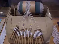 Na zdjęciu widać 24 nawleczone klocki – drewniane wałeczki, szersze na dole, a węższe o góry. Wąskie części klocków owinięte są nićmi biegnącymi ku widocznej na dalszym planie, częściowo wykonanej koronki, przypiętej do wypchanej poduszki w kształcie wałka. Obok leży fragment ukończonej już koronki o tym samym wzorze.