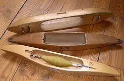 Trzy czółenka tkackie leżące na drewnianej podłodze. Najbliżej patrzącego widać współczesne czółenko z dnem, zawierające nawiniętą cewkę z zieloną przędzą. Za nim znajduje się starsze, puste czółenko z dnem. Za nim natomiast leży na boku puste czółenko bez dna, również już nieco zużyte.