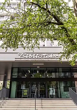 Przeszklone wejście do budynku. Nad wejściem napis Biblioteka Wojewódzka. Na pierwszym planie gałąź drzewa.