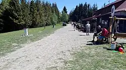 Zdjęcie wykonane w słoneczny, letni dzień przedstawia piaszczystą drogę sfotografowaną z perspektywy idącego nią człowieka. Po prawej stronie wzdłuż drogi jest niski budynek, przed nim stoliki i ławeczki na których siedzą ludzie. Po lewej stronie wzdłuż drogi rośnie trawa, zaraz za nią zaczyna się las.