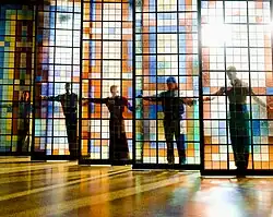 na zdjęciu widać szklany witraż wysoki od podłogi do sufitu, zbudowany z kwadratowych kolorowych szkieł, 6 części, za każdą stoi tancerz obejmuje część witraża rękoma