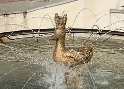 Zdjęcie przedstawia okrągłą fontannę, pośrodku której stoi figura kaczki w koronie na głowie.