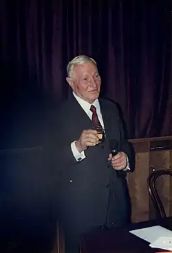 Starszy mężczyzna z siwymi włosami, ubrany w garnitur z czerwonym krawatem. W prawej ręce trzyma kieliszek, a w lewej mikrofon.