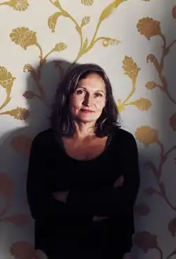 Kolorowe zdjęcie kobiety od pasa w górę. Ma założone ręce i lekko się uśmiecha, a jej rozpuszczone włosy sięgają ramion. Jest oparta o ścianę ze złotym wzorem roślinnym.